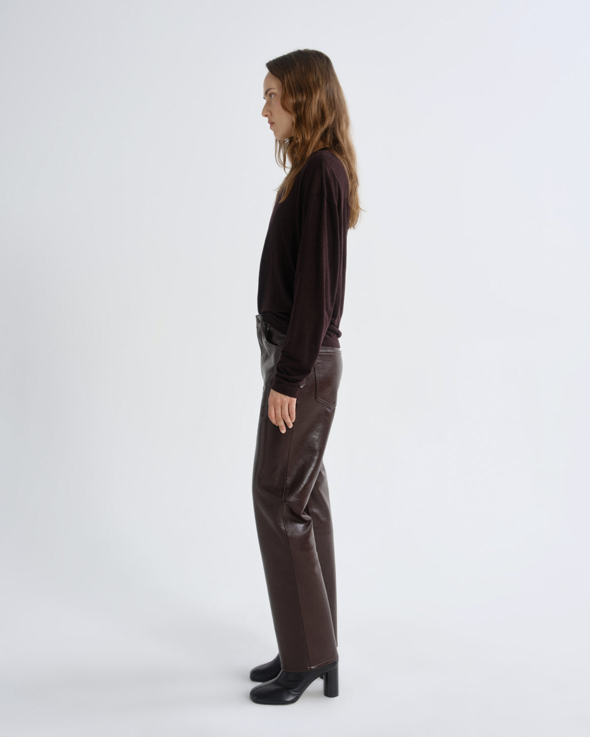 A woman stands in profile wearing a dark brown long-sleeve top, dark brown wide-leg pants, and black heeled boots against a plain light gray studio background.