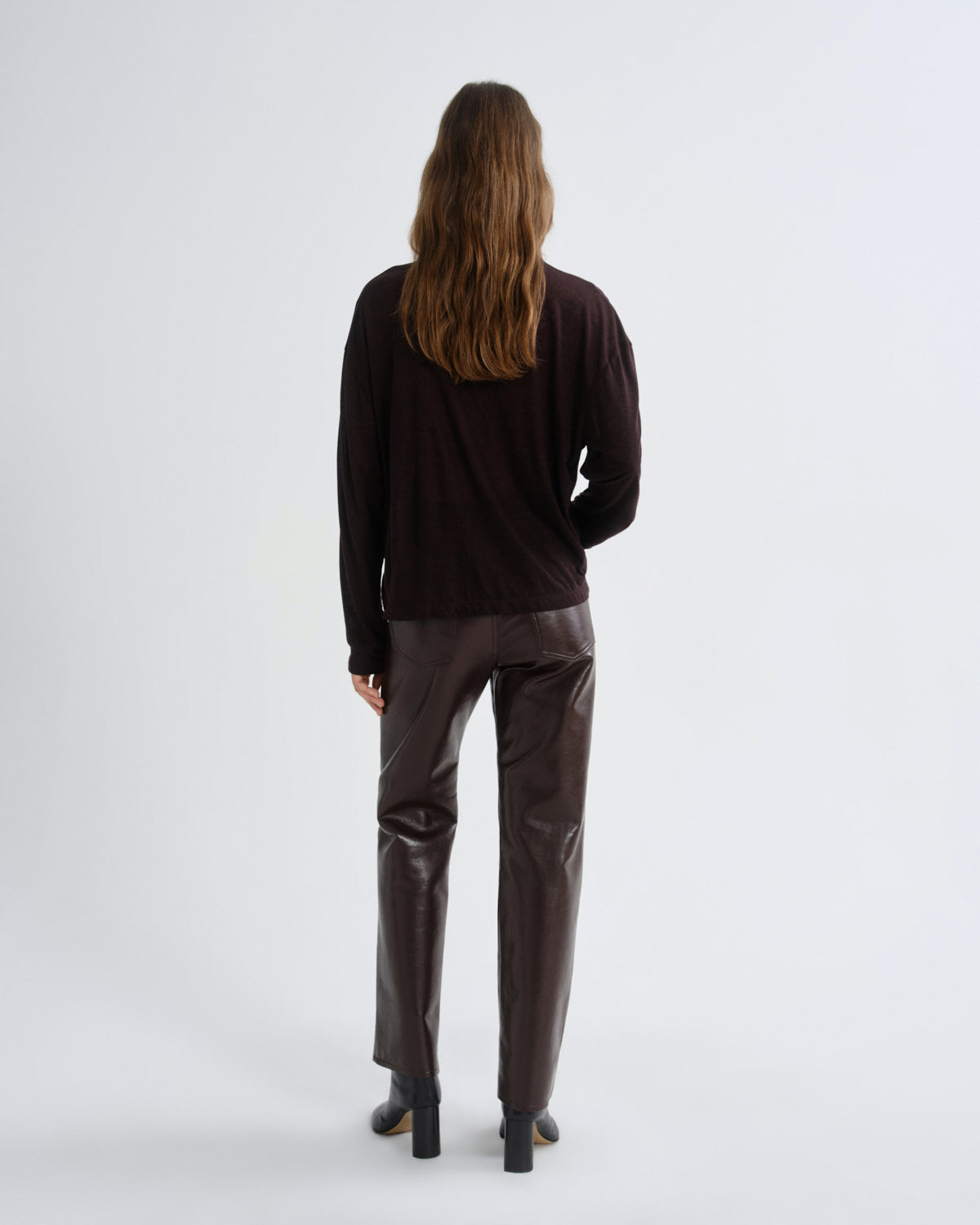 A woman is shown from the back wearing a dark brown long-sleeve top and dark brown pants, standing against a plain light gray studio background.