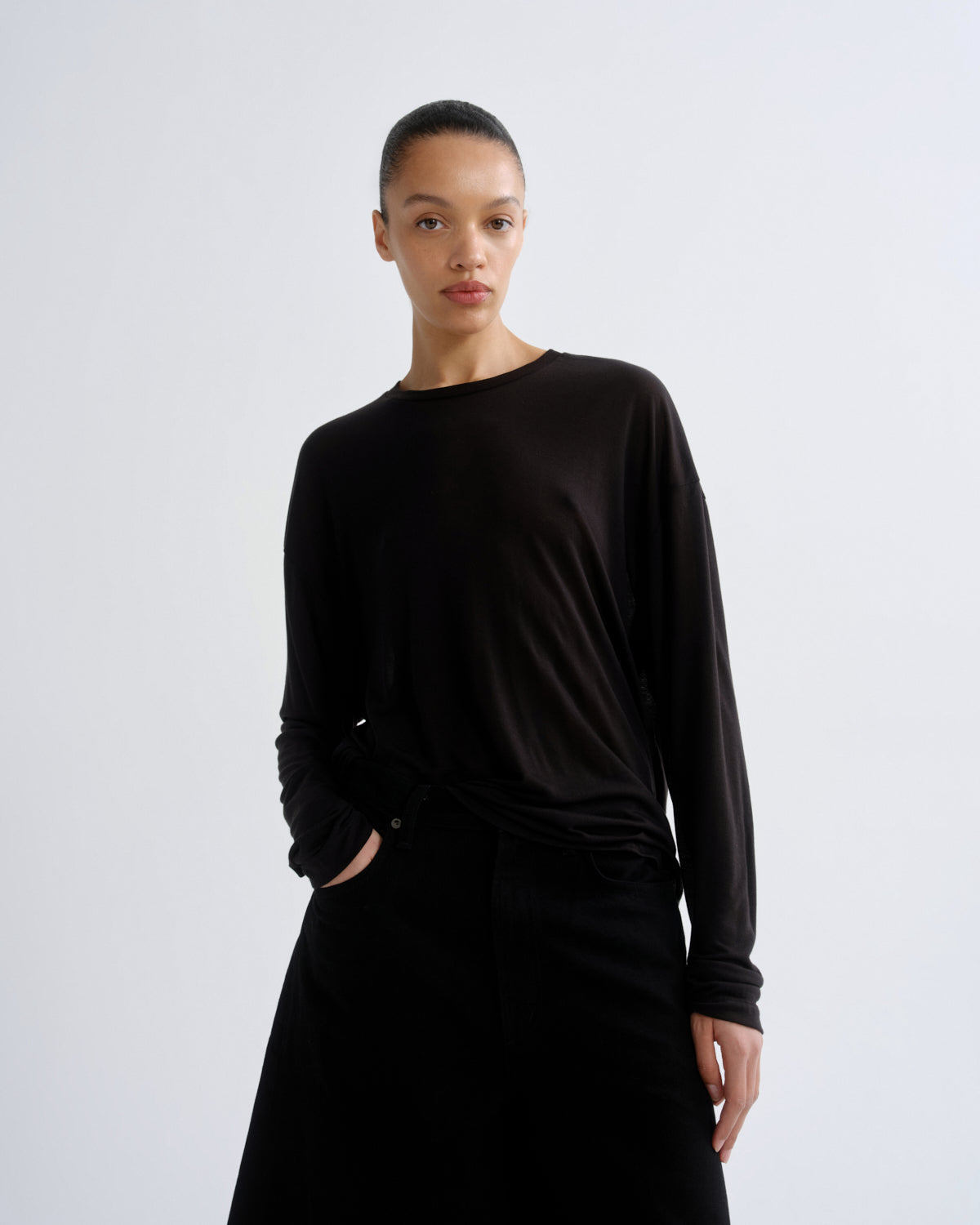 A model wearing a black long-sleeve knit top and black skirt, standing against a light gray studio background.