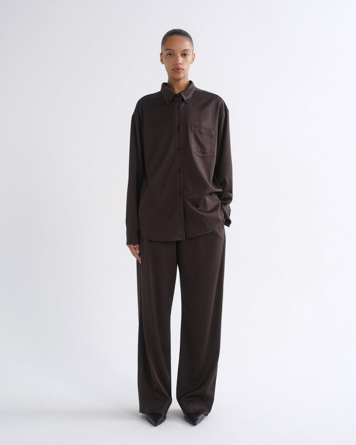 A model stands against a light gray studio background wearing a dark brown long-sleeve button-up shirt and matching dark brown wide-leg trousers.