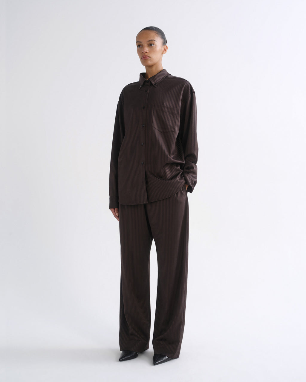 A model stands against a plain light gray studio background wearing a dark brown long-sleeve button-up shirt and matching dark brown wide-leg trousers.