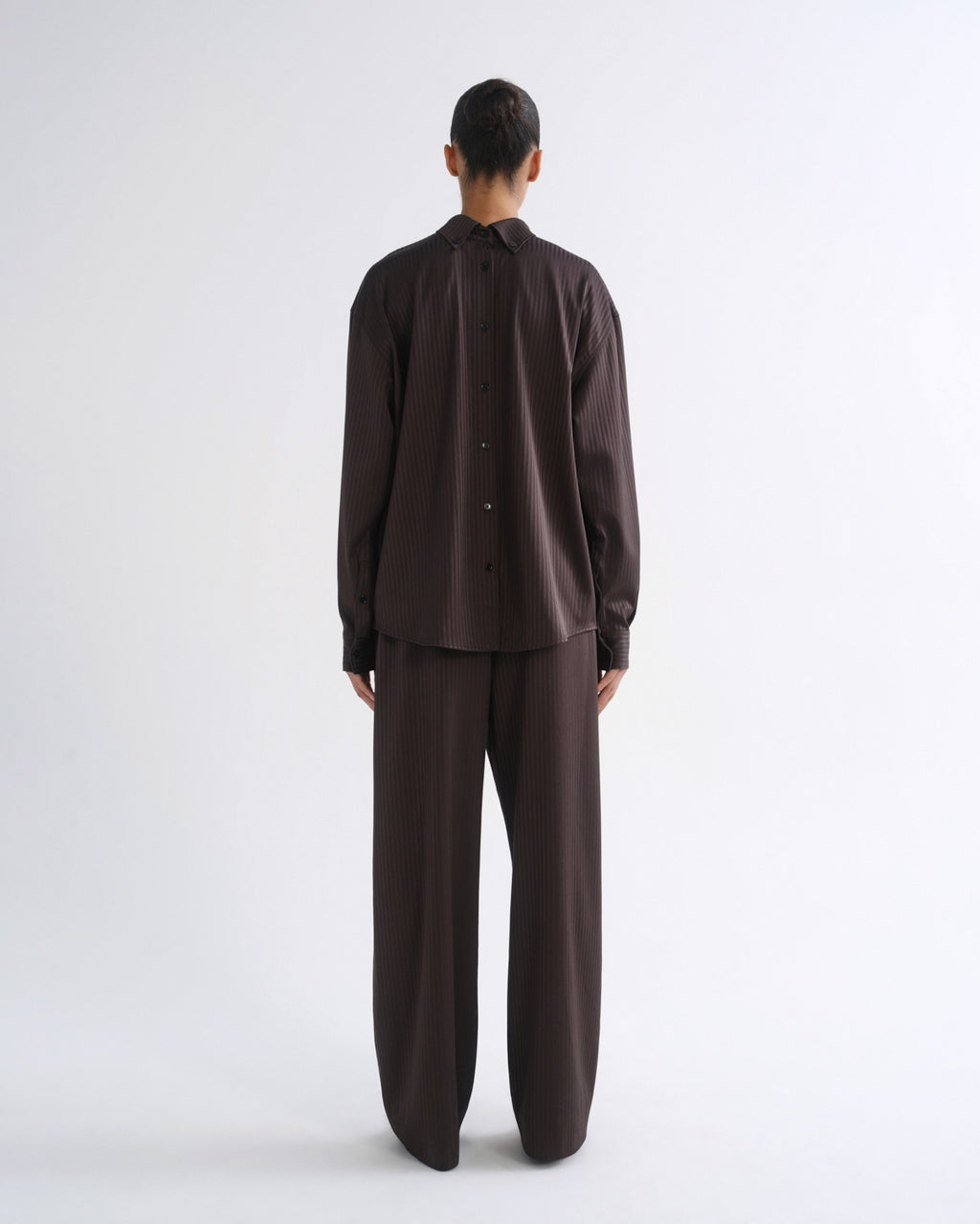 A model viewed from the back wearing a dark brown button-up shirt and matching wide-leg trousers against a plain white studio background.