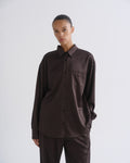 A model wearing a dark brown ribbed long-sleeve button-up shirt and matching dark brown trousers, photographed against a neutral light gray studio background.