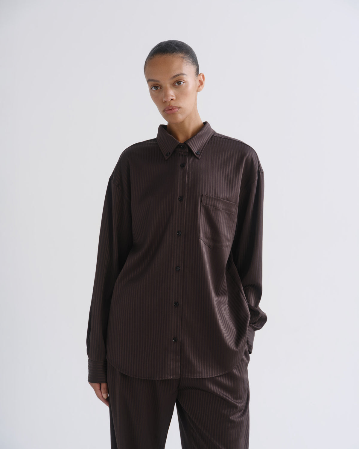 A model wearing a dark brown ribbed long-sleeve button-up shirt and matching dark brown trousers, photographed against a neutral light gray studio background.