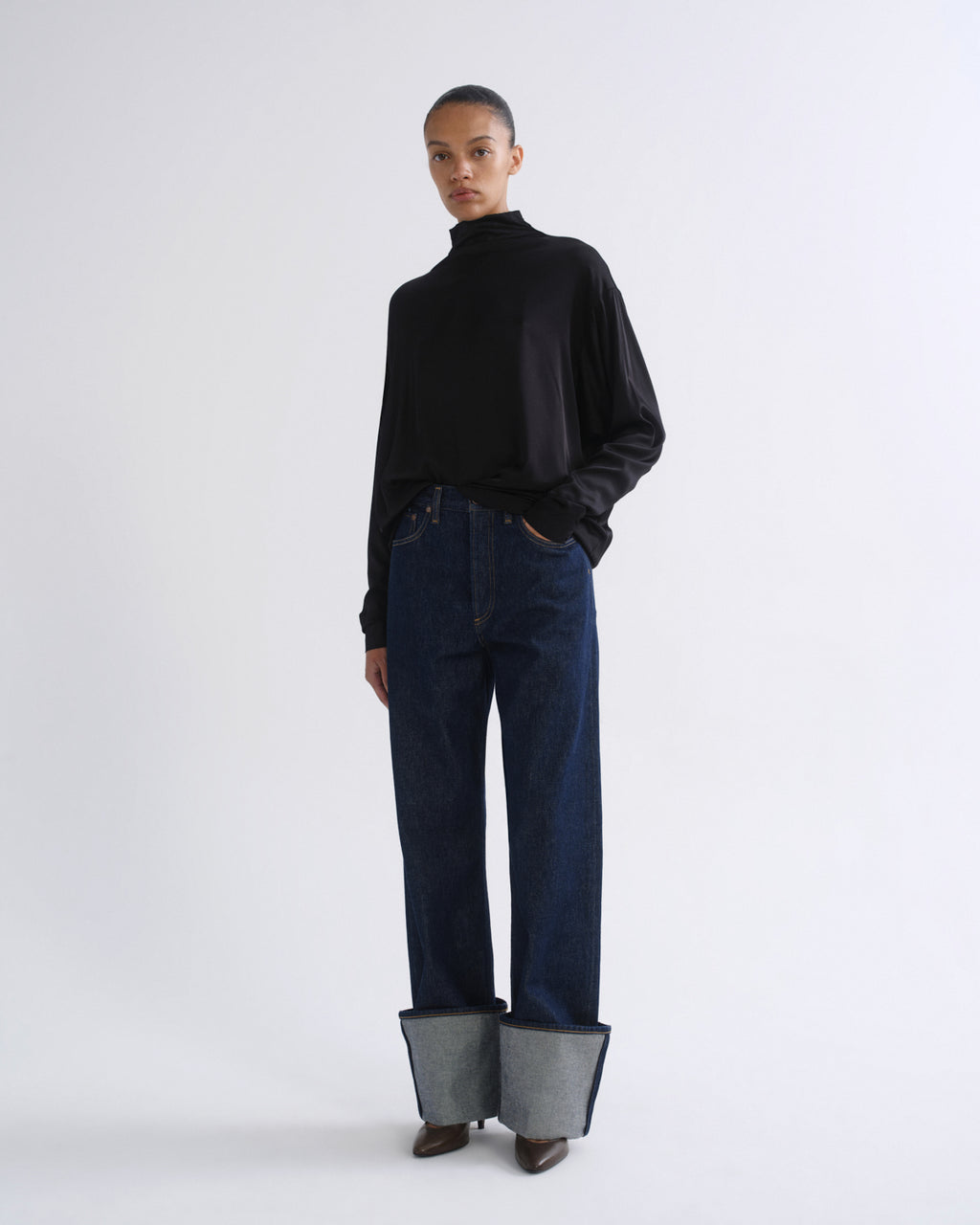 Woman wearing a black long sleeve mock neck top with dark indigo jeans on a grey background.
