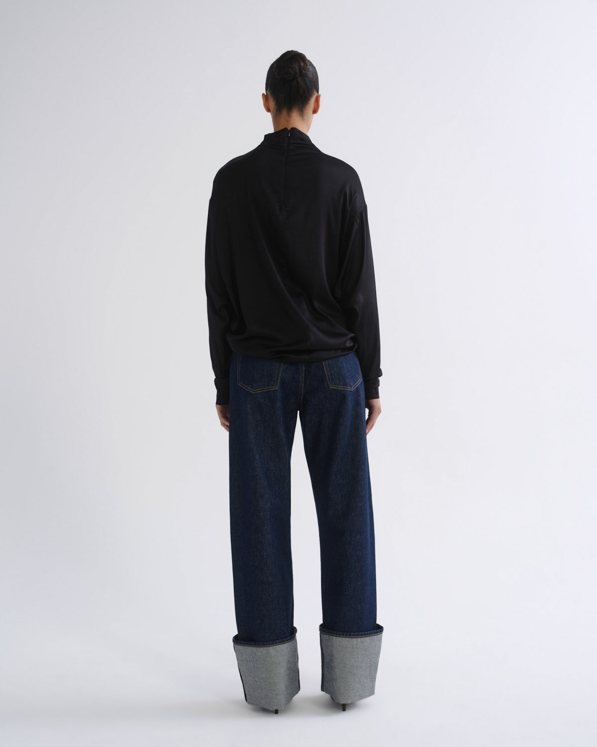 Woman wearing a black long sleeve mock neck top with dark indigo jeans viewed from the back on a grey background.