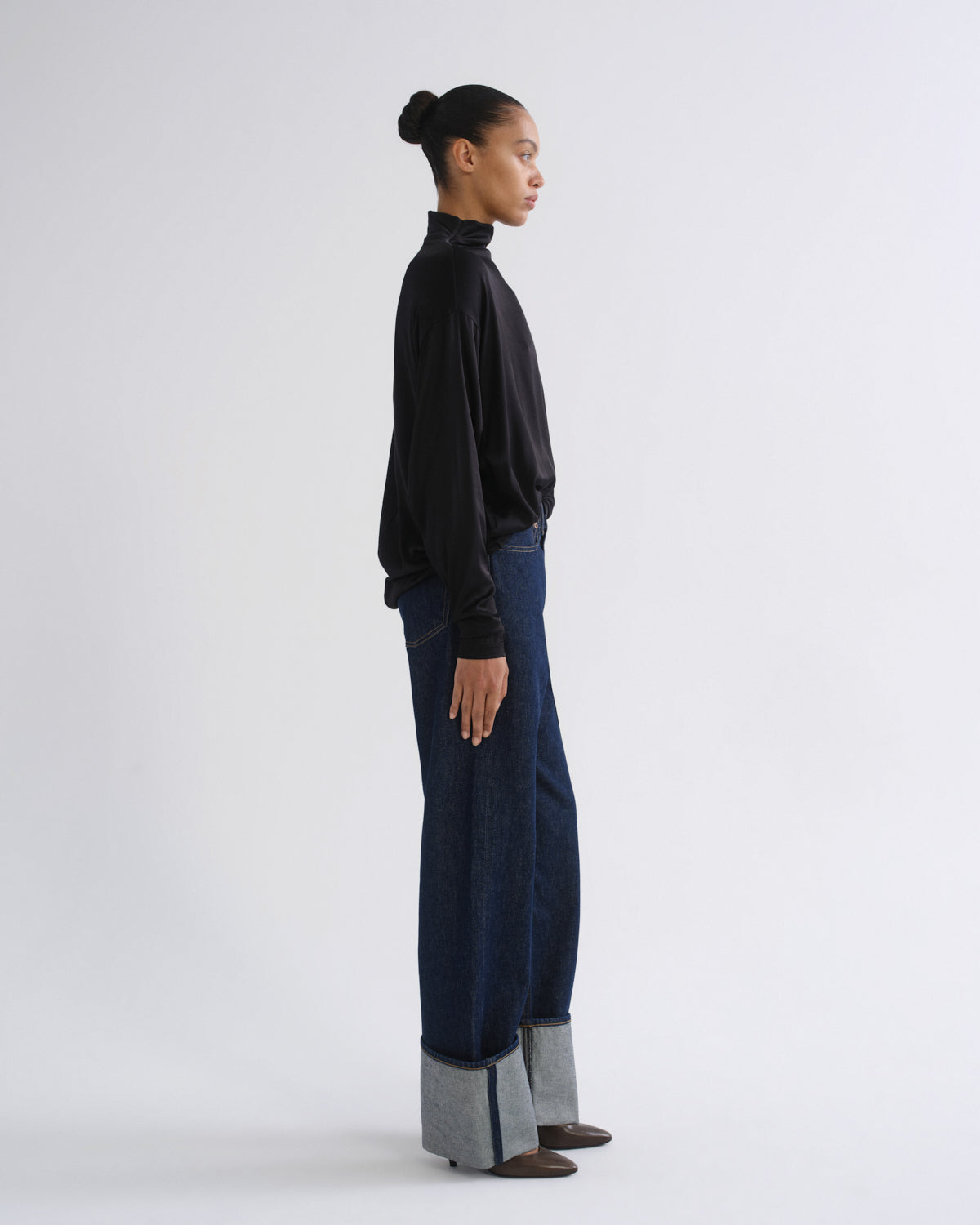 Woman wearing a black long sleeve mock neck top with dark indigo jeans viewed from the side on a grey background.