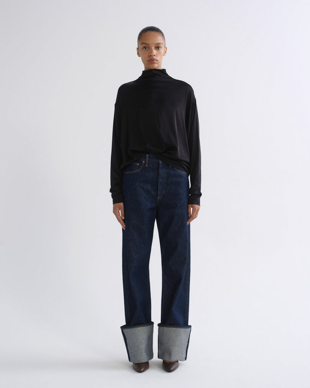Woman wearing a black long sleeve mock neck top with dark indigo jeans on a grey background.