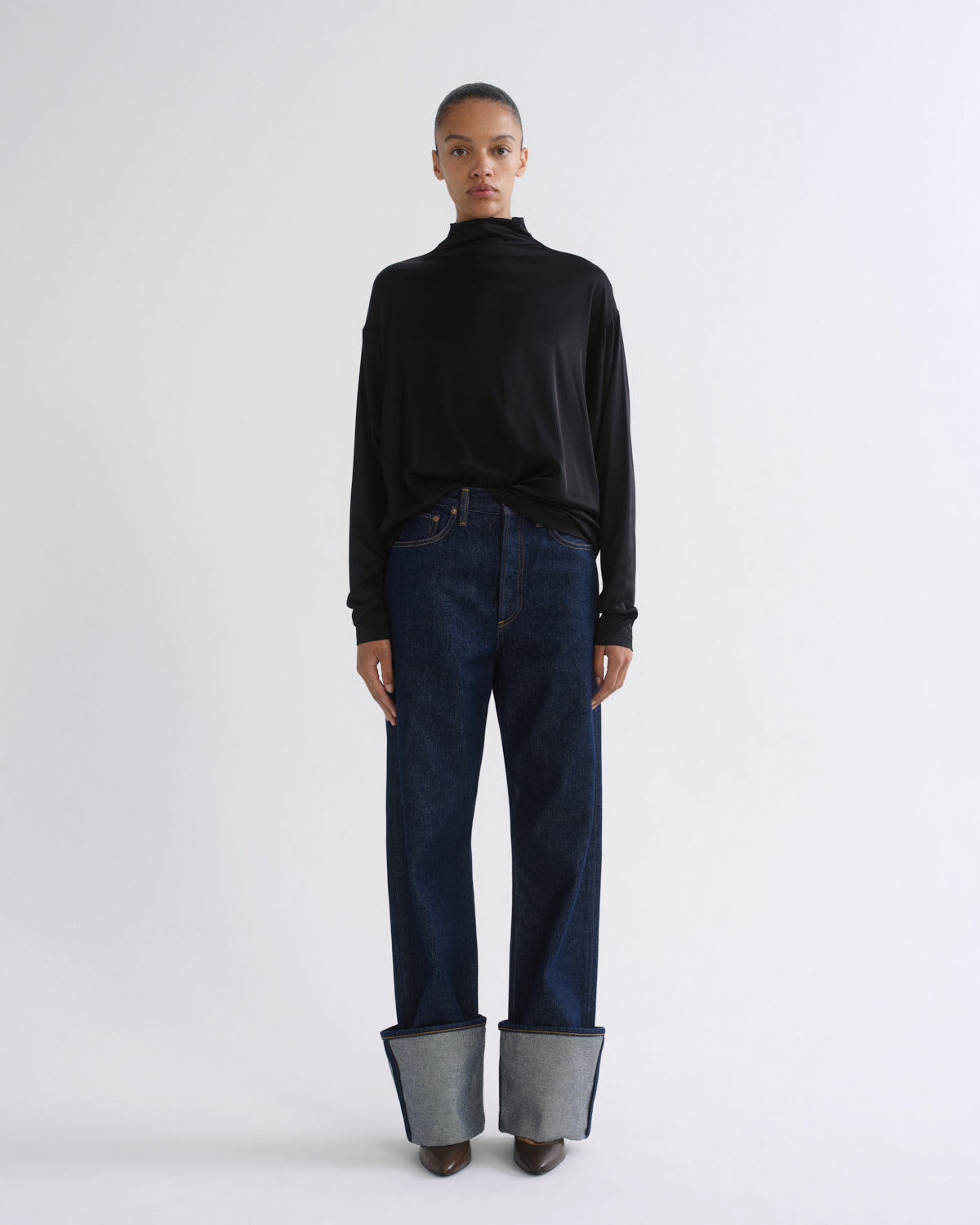 Woman wearing a black long sleeve mock neck top with dark indigo jeans on a grey background.