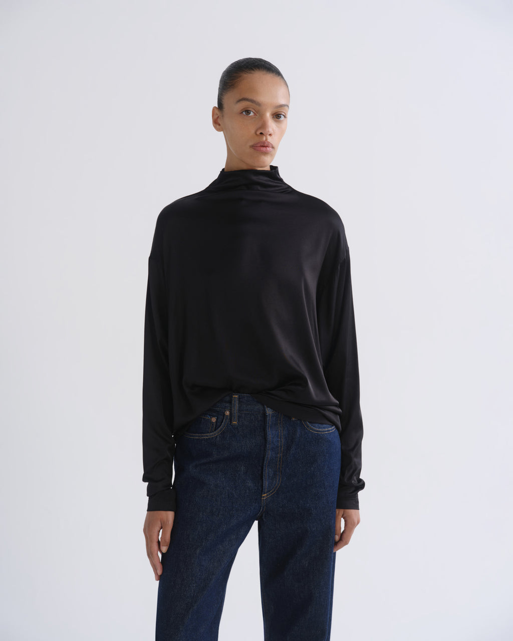 Woman wearing a black long sleeve mock neck top with dark indigo jeans on a grey background.