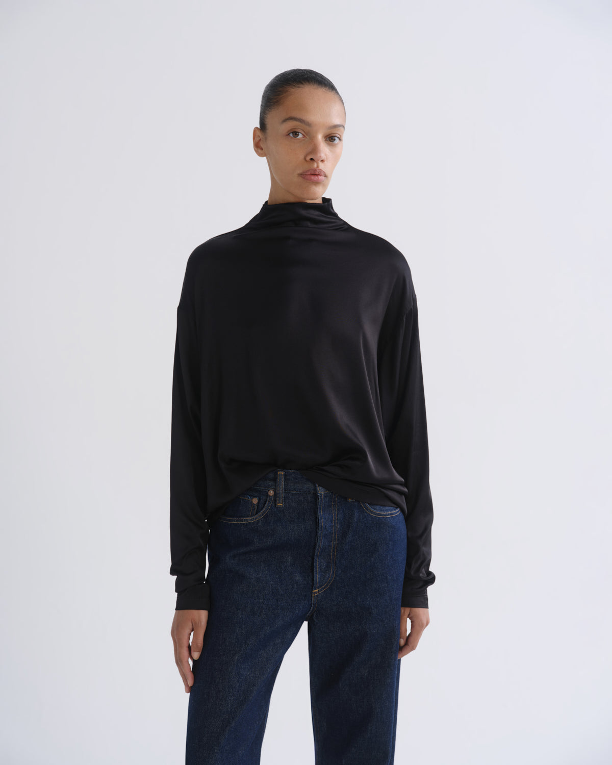 Woman wearing a black long sleeve mock neck top with dark indigo jeans on a grey background.