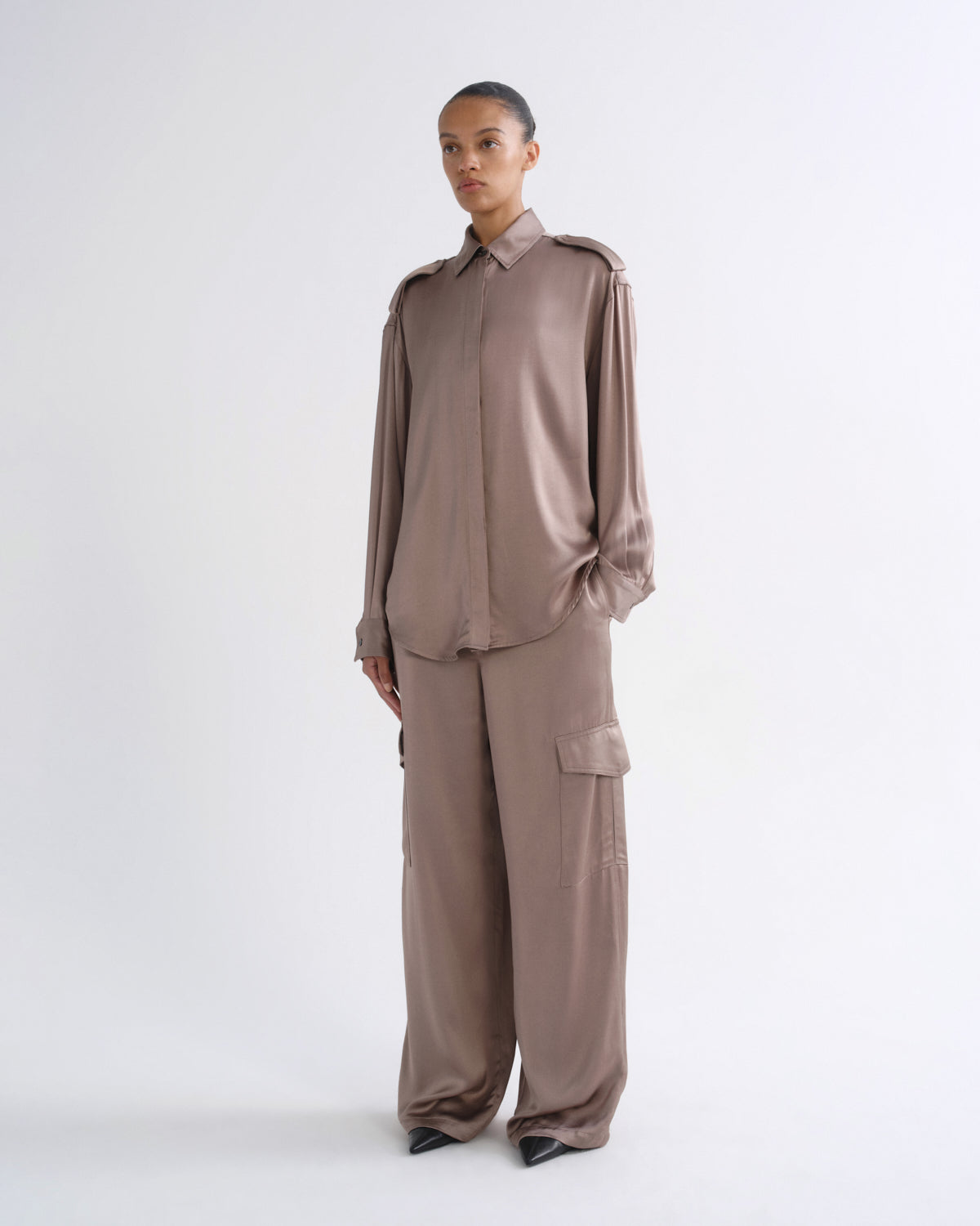 A model stands against a light gray studio background wearing a matching taupe-brown two-piece outfit consisting of a long-sleeve button-front shirt with shoulder epaulets and wide-leg cargo pants.