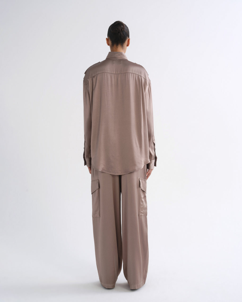 A model wearing a long-sleeve, collared button-up shirt and matching cargo pants in a muted taupe-brown color, shot against a plain light gray studio background.
