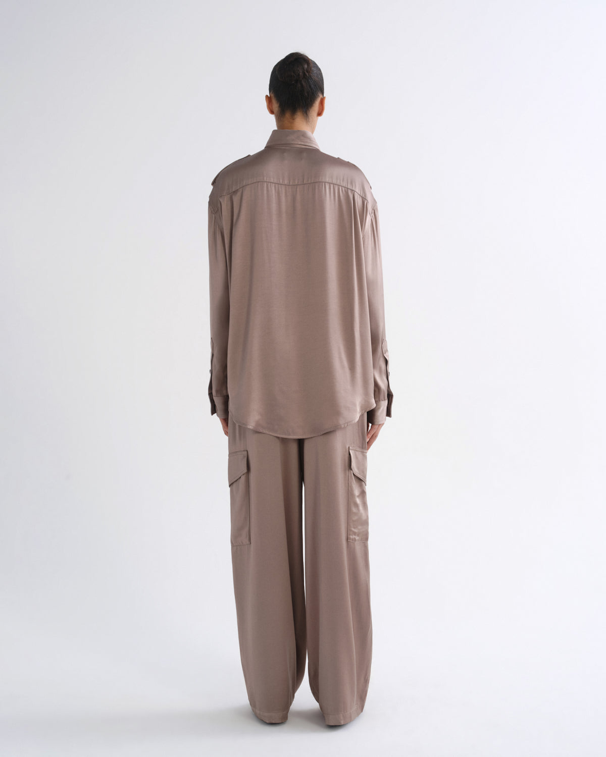 A model wearing a long-sleeve, collared button-up shirt and matching cargo pants in a muted taupe-brown color, shot against a plain light gray studio background.
