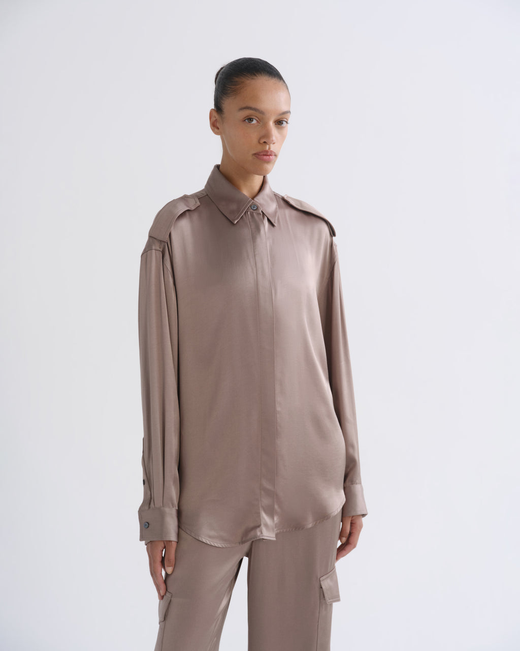 A model wearing a long-sleeve, collared button-up shirt and matching cargo pants in a muted taupe-brown color, shot against a plain light gray studio background.