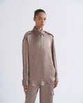 A model wearing a long-sleeve, collared button-up shirt and matching cargo pants in a muted taupe-brown color, shot against a plain light gray studio background.
