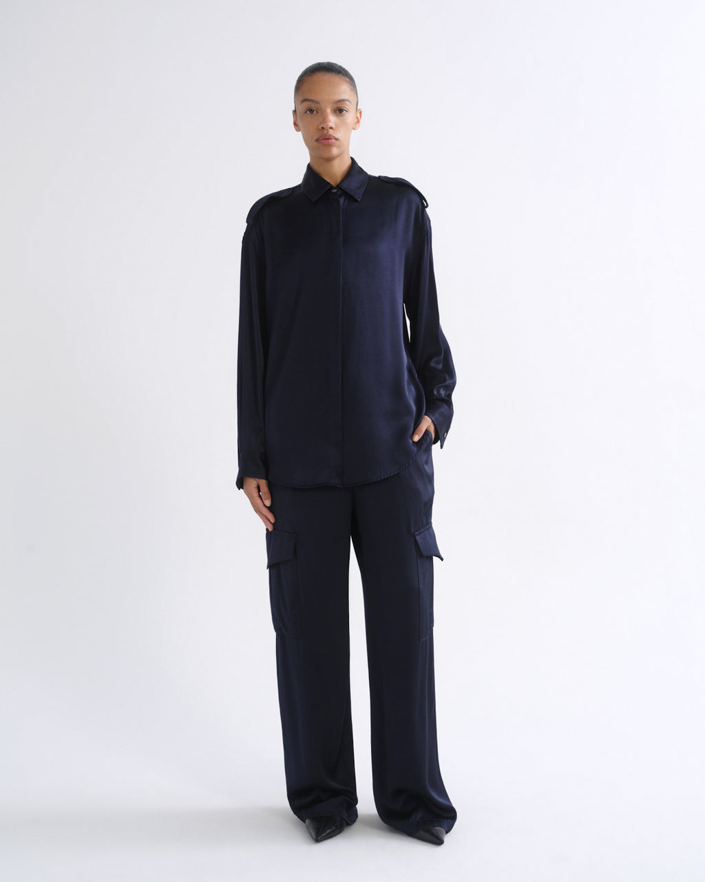 A model stands against a plain white studio background wearing a dark navy blue long-sleeve collared shirt and matching wide-leg cargo pants.