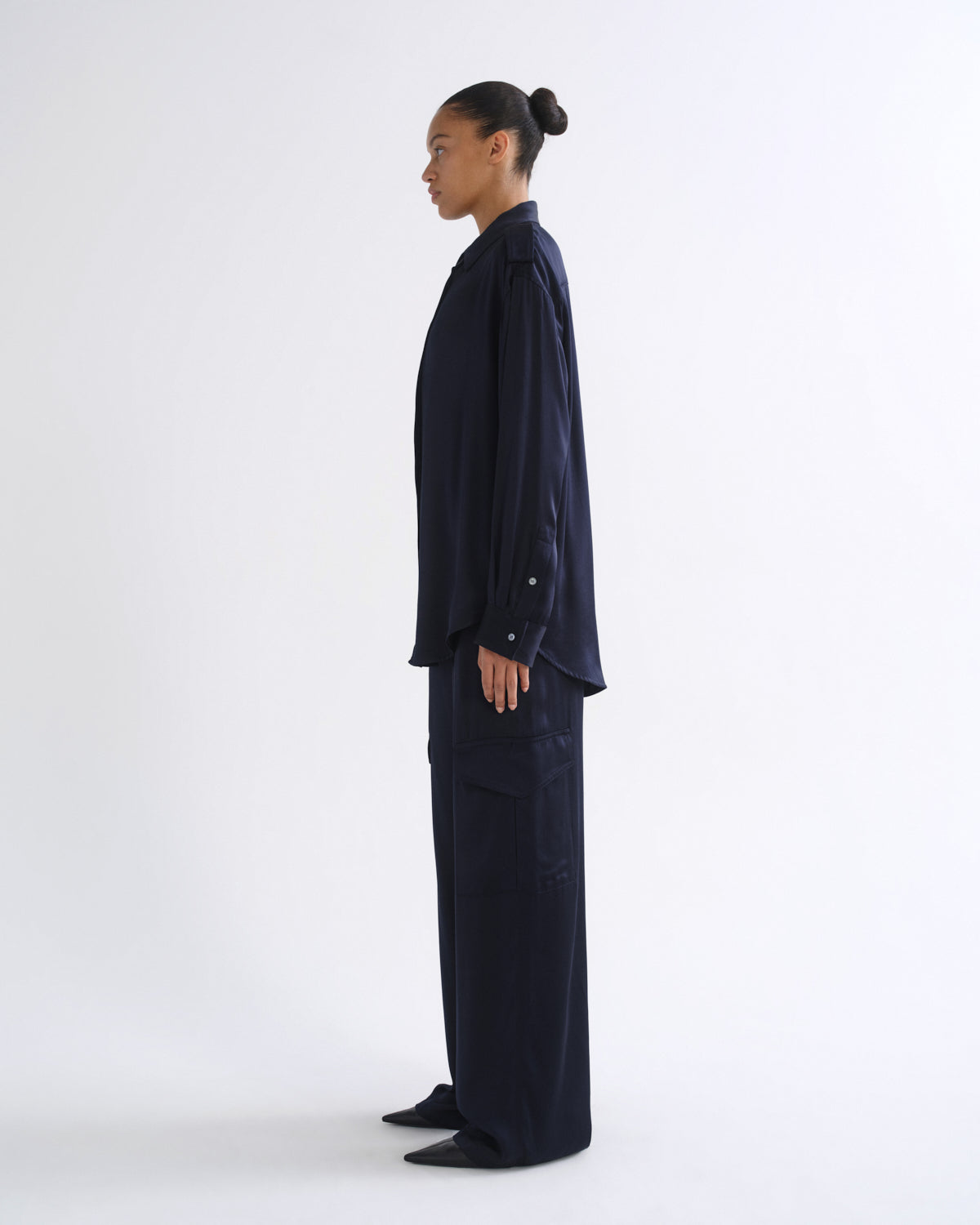 A model in side profile wearing a dark navy blue long-sleeve button-up shirt and matching wide-leg trousers, standing against a plain white studio background.
