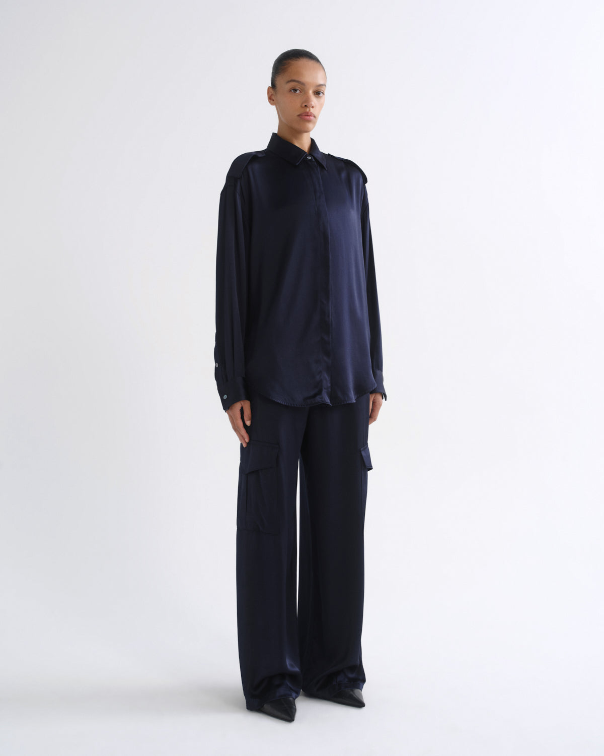 A model stands against a plain white studio background wearing a dark navy blue satin long-sleeve button-up shirt and matching wide-leg trousers.