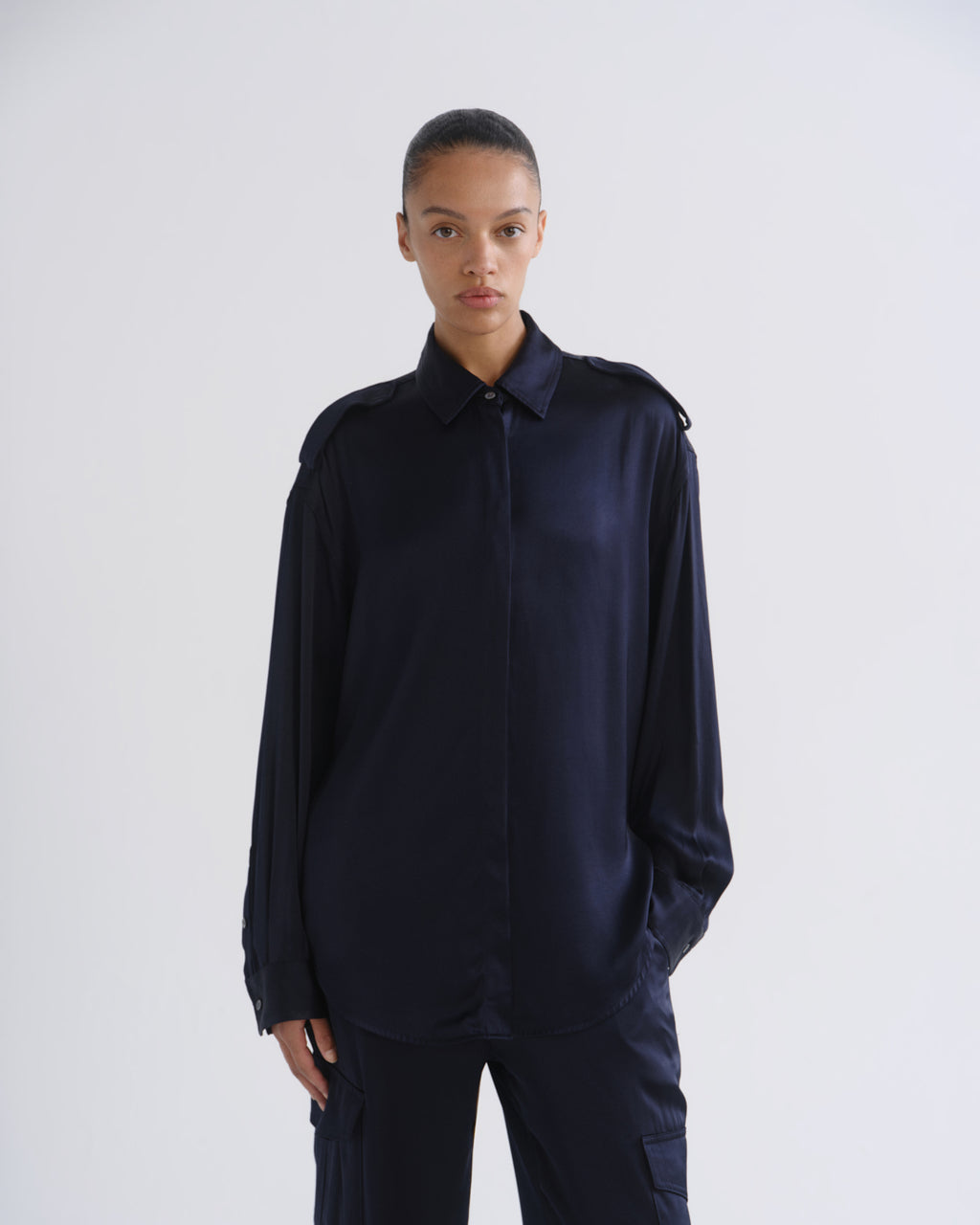 A model wearing a dark navy blue satin long-sleeve shirt with shoulder epaulets and matching dark trousers, standing against a light gray studio background.