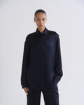 A model wearing a dark navy blue satin long-sleeve shirt with shoulder epaulets and matching dark trousers, standing against a light gray studio background.