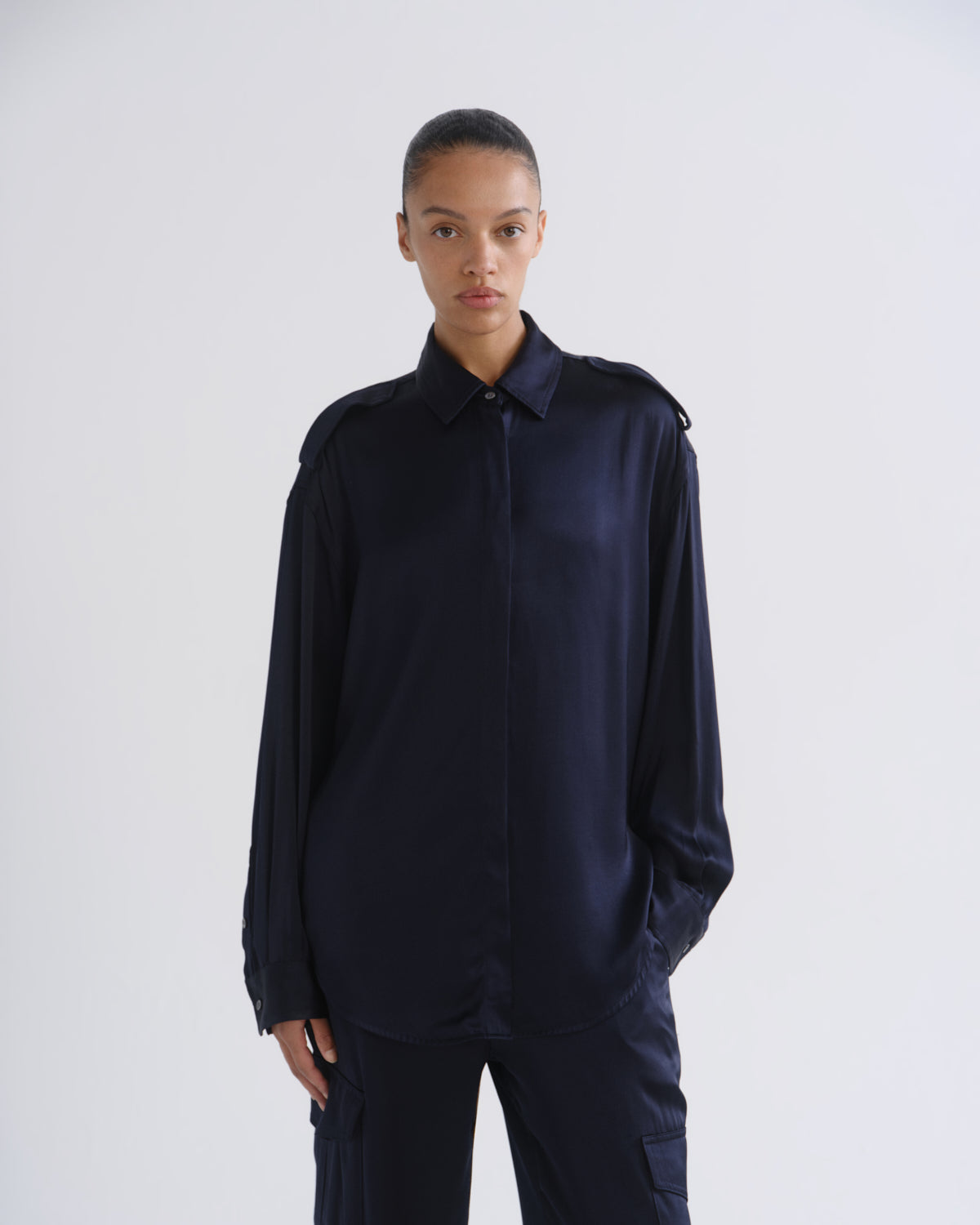A model wearing a dark navy blue satin long-sleeve shirt with shoulder epaulets and matching dark trousers, standing against a light gray studio background.