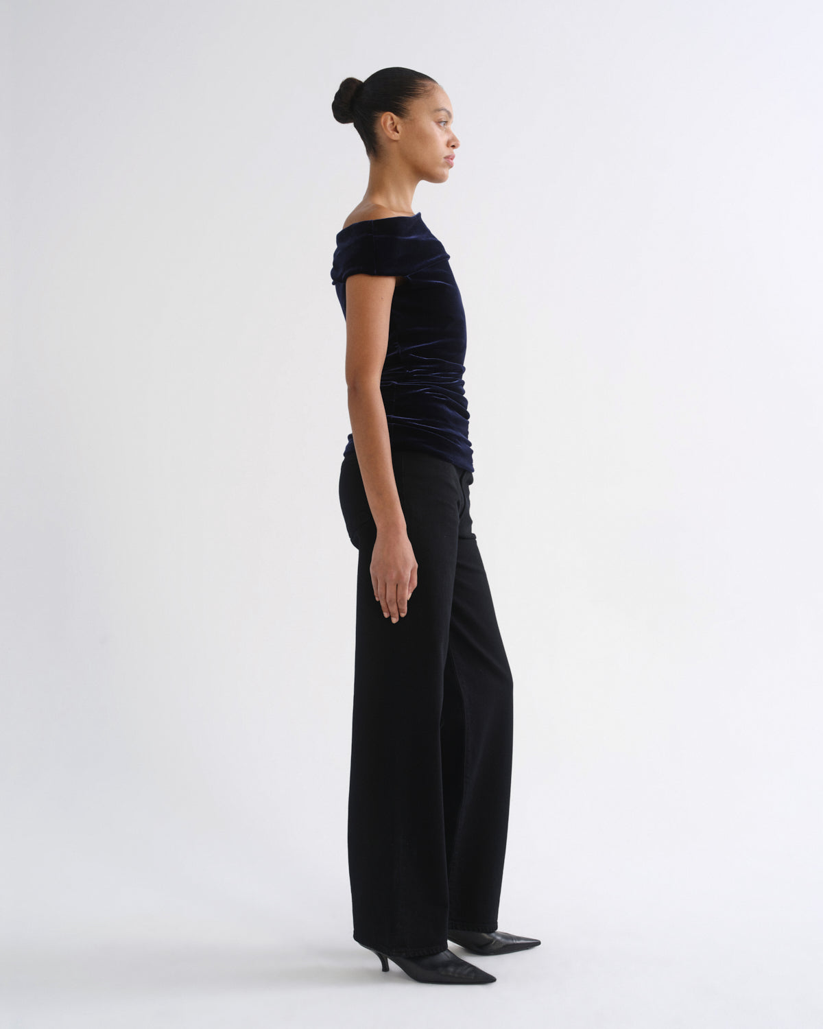 Woman wearing a black sleeveless draped top with black straight leg pants viewed from the side on a grey background.