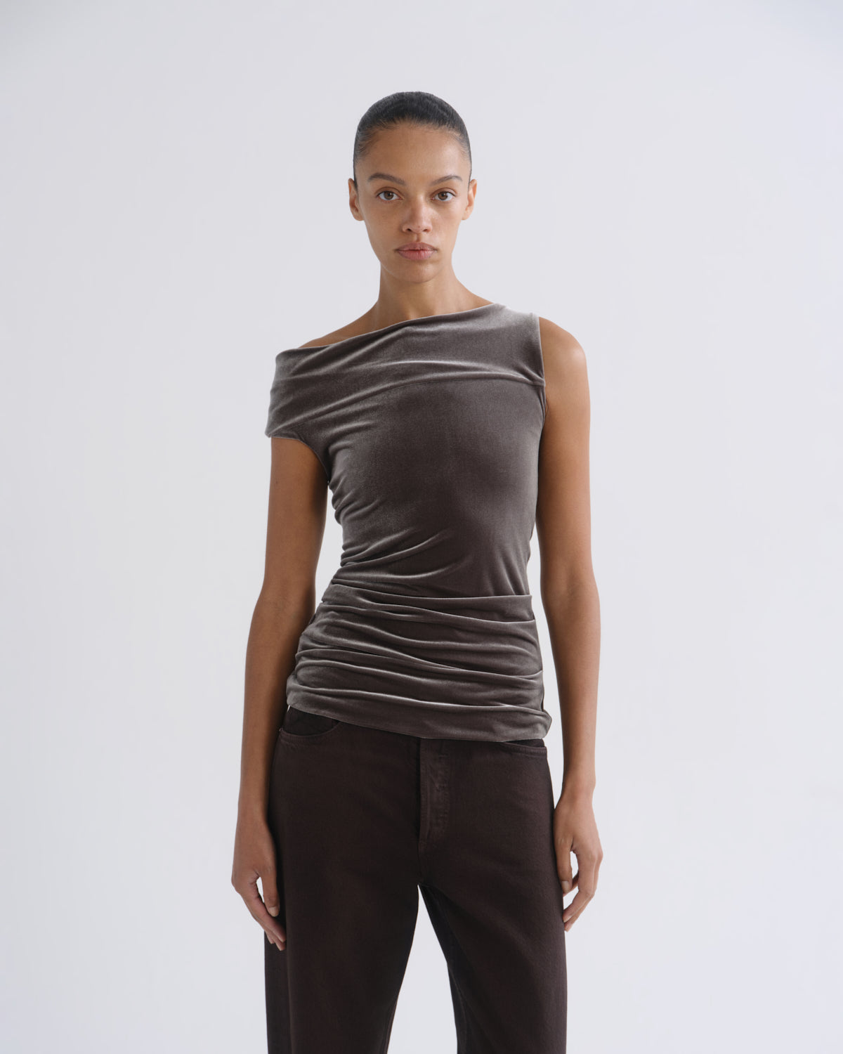 Woman wearing a brown sleeveless draped top with dark brown wide leg pants on a grey background.