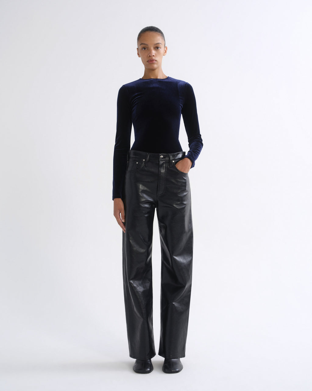 A model stands against a plain light gray studio background wearing a dark navy long-sleeve velvet top and shiny black wide-leg pants.
