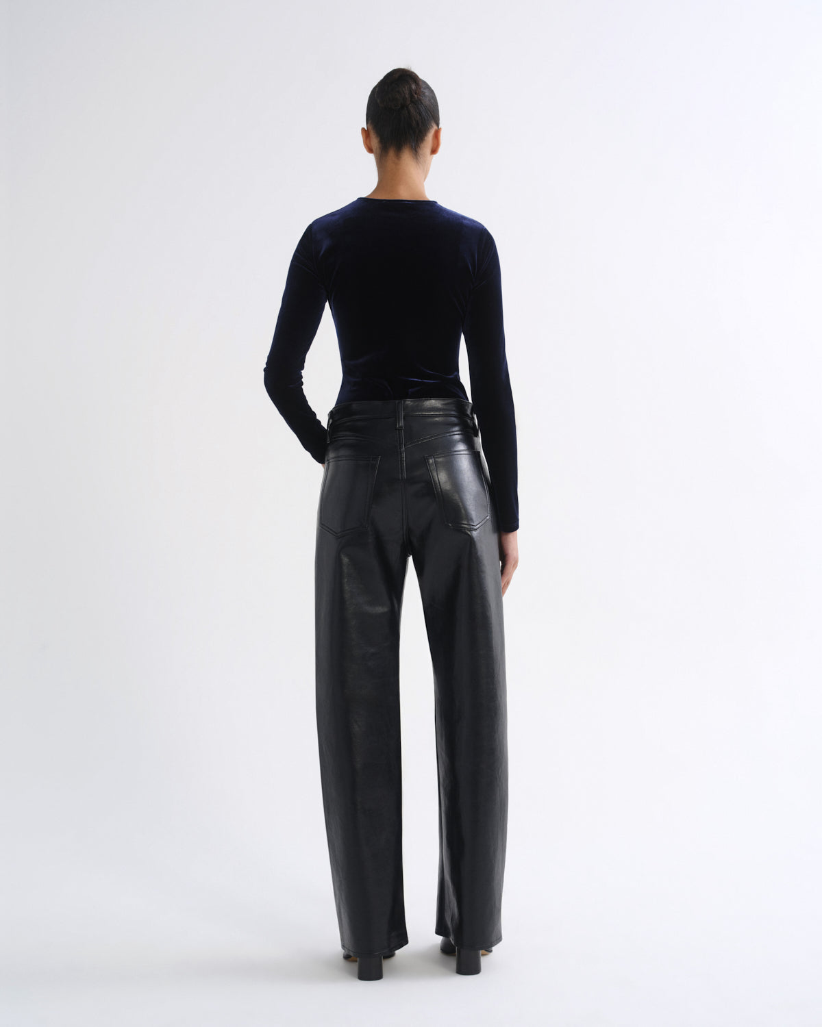 A model shown from the back wearing a black velvet long-sleeve top and black wide-leg leather trousers against a plain white studio background.