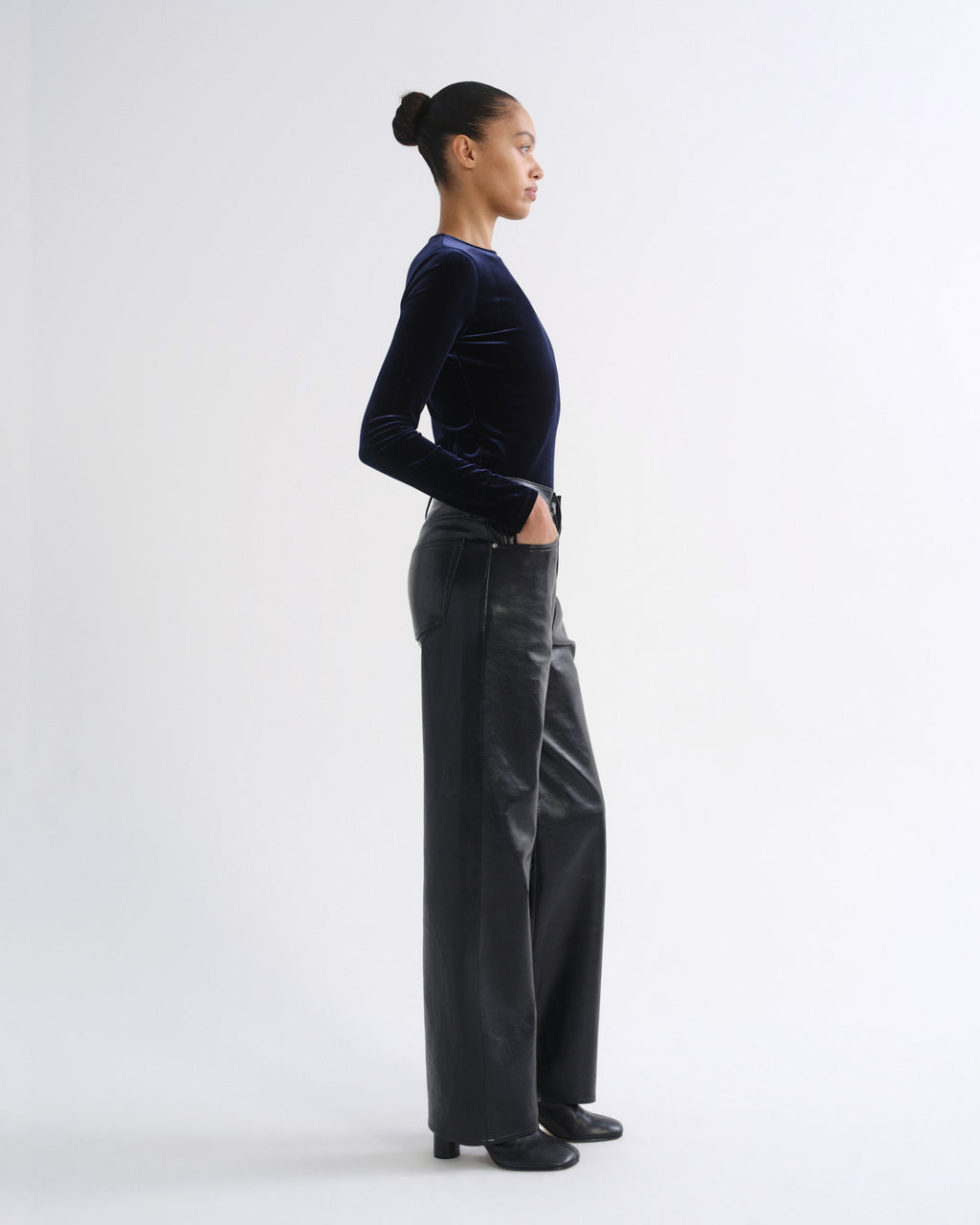 A model in profile wearing a dark navy velvet long-sleeve top and dark gray wide-leg trousers, standing against a plain light gray studio background.