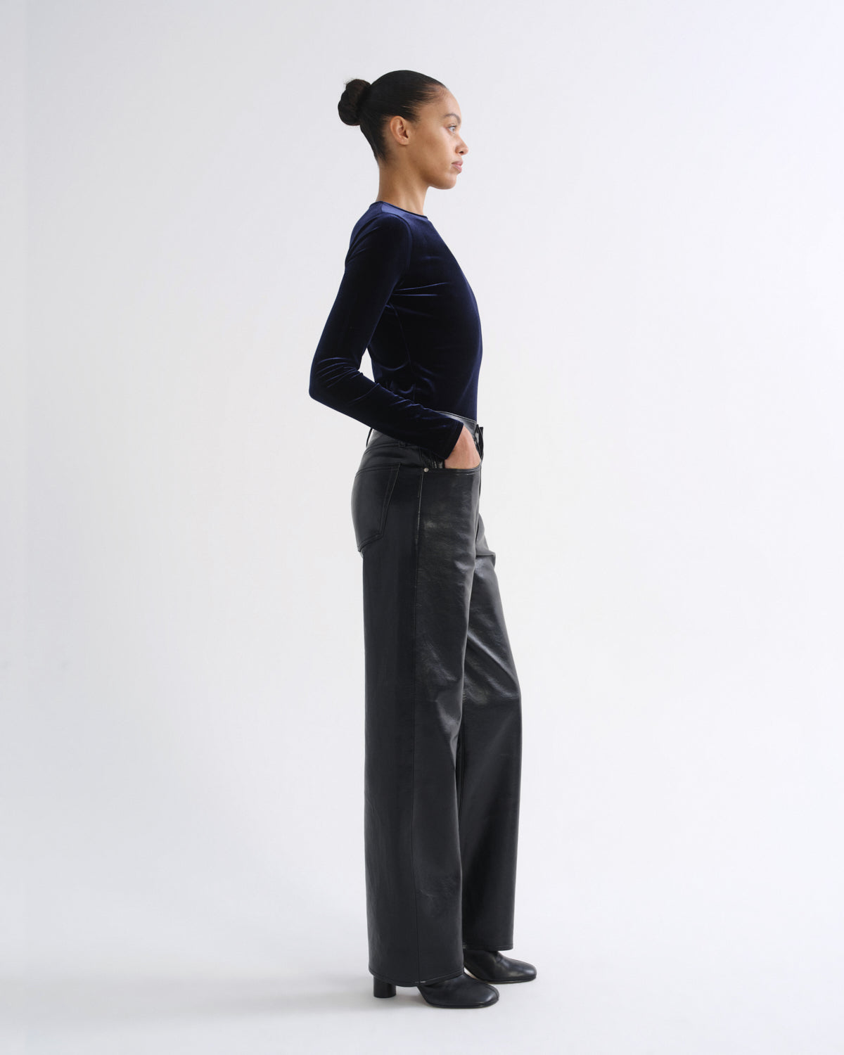A model in profile wearing a dark navy velvet long-sleeve top and dark gray wide-leg trousers, standing against a plain light gray studio background.