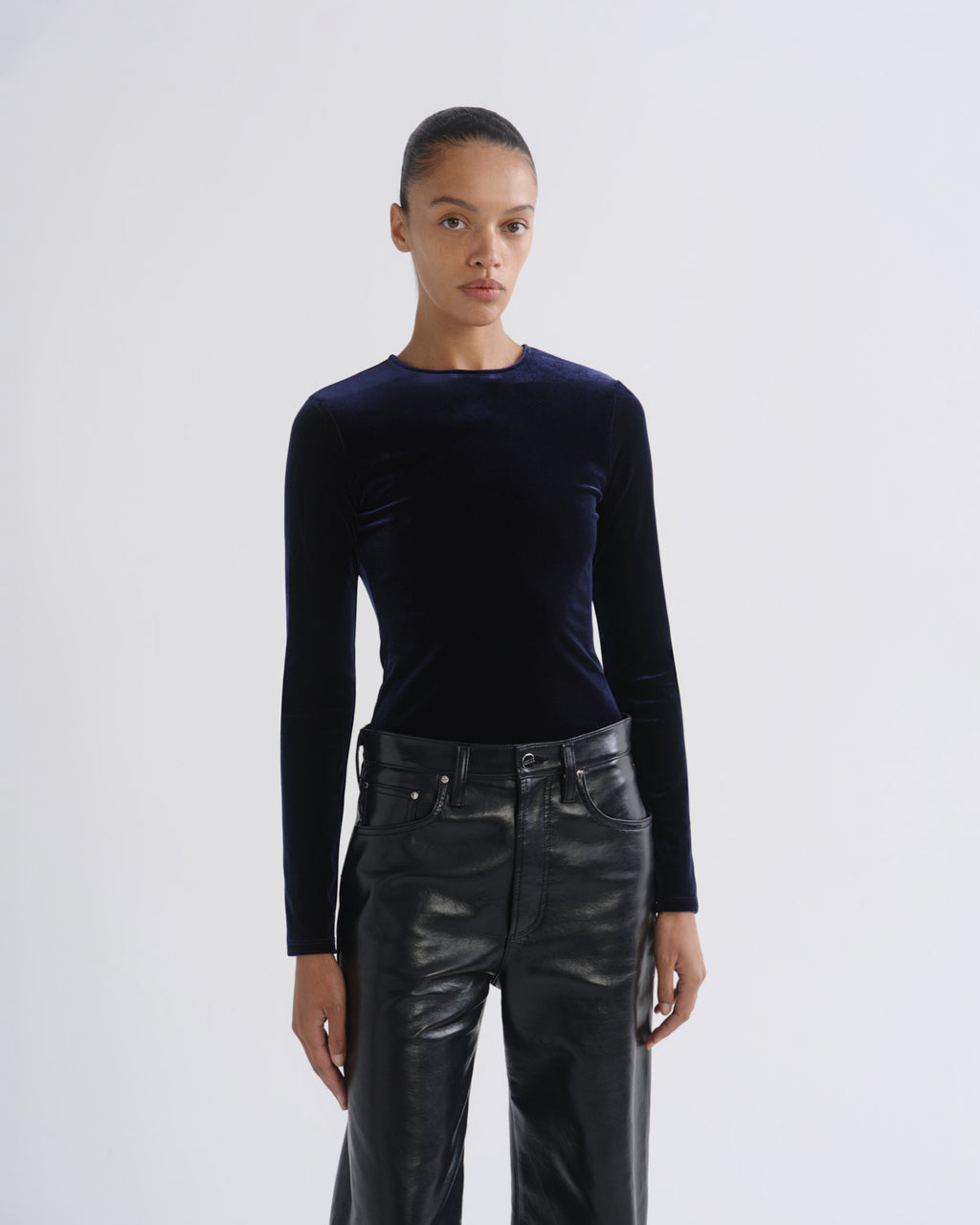 A model wearing a dark navy blue long-sleeve velvet top and black leather pants, standing against a light gray studio background.
