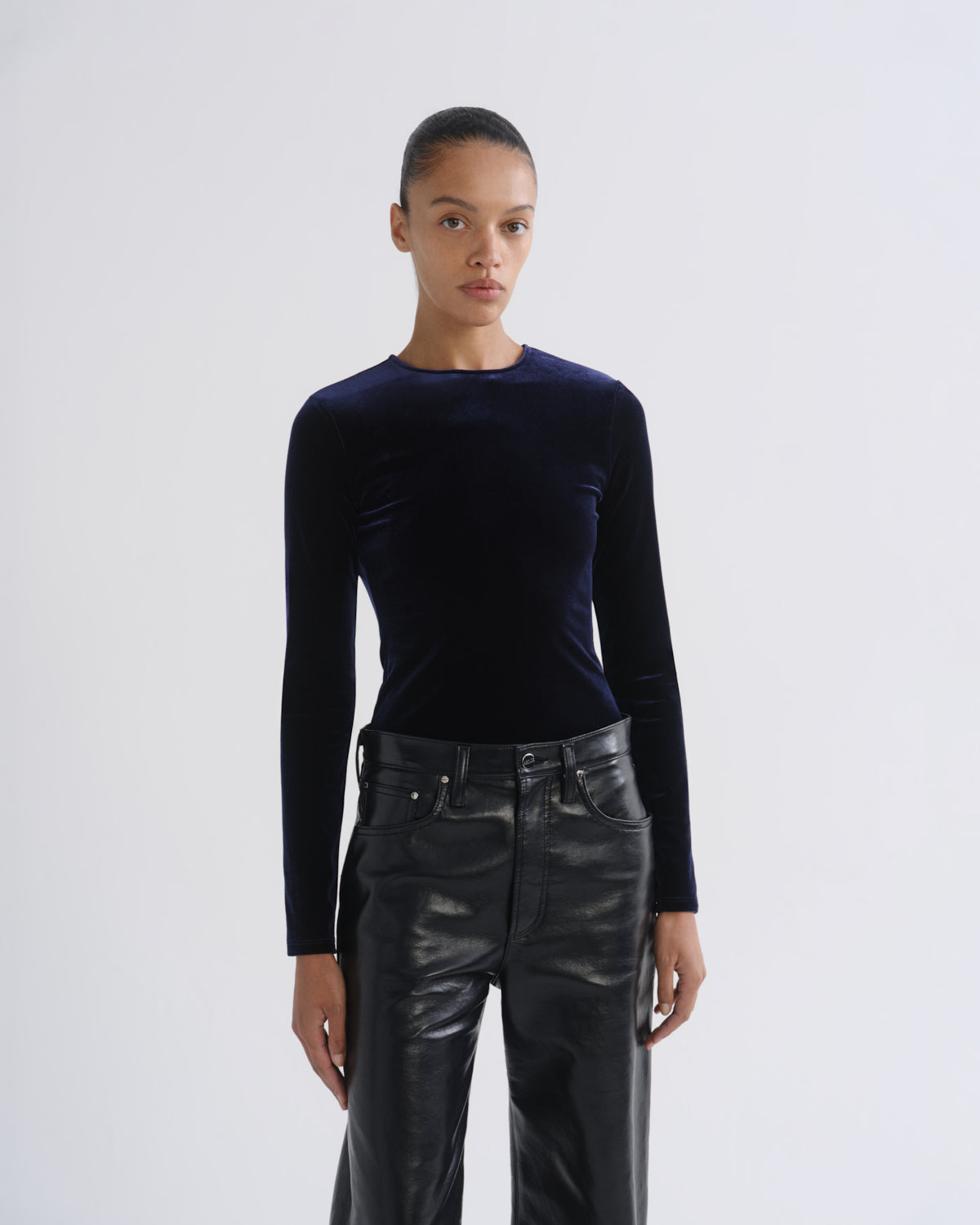 A model wearing a dark navy blue long-sleeve velvet top and black leather pants, standing against a light gray studio background.