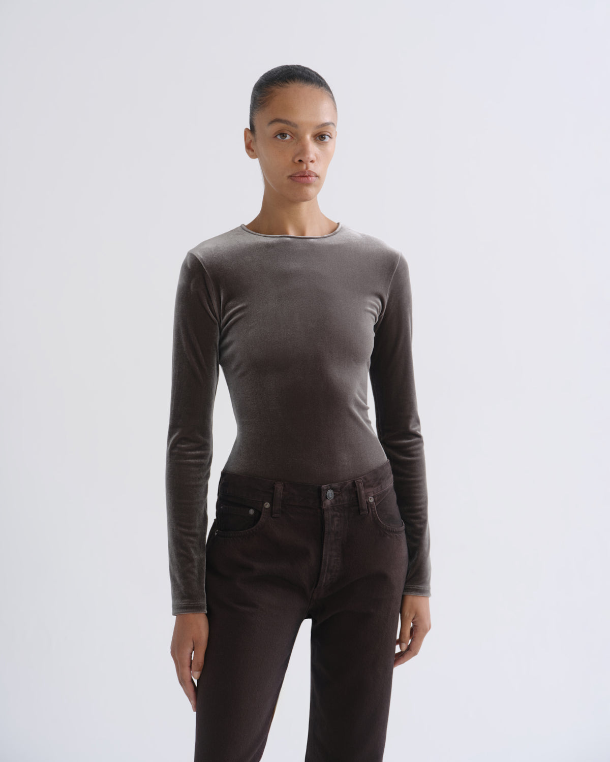 A model wearing a fitted, long-sleeve crewneck top in a dark taupe-brown color, paired with dark brown pants, against a plain light gray studio background.