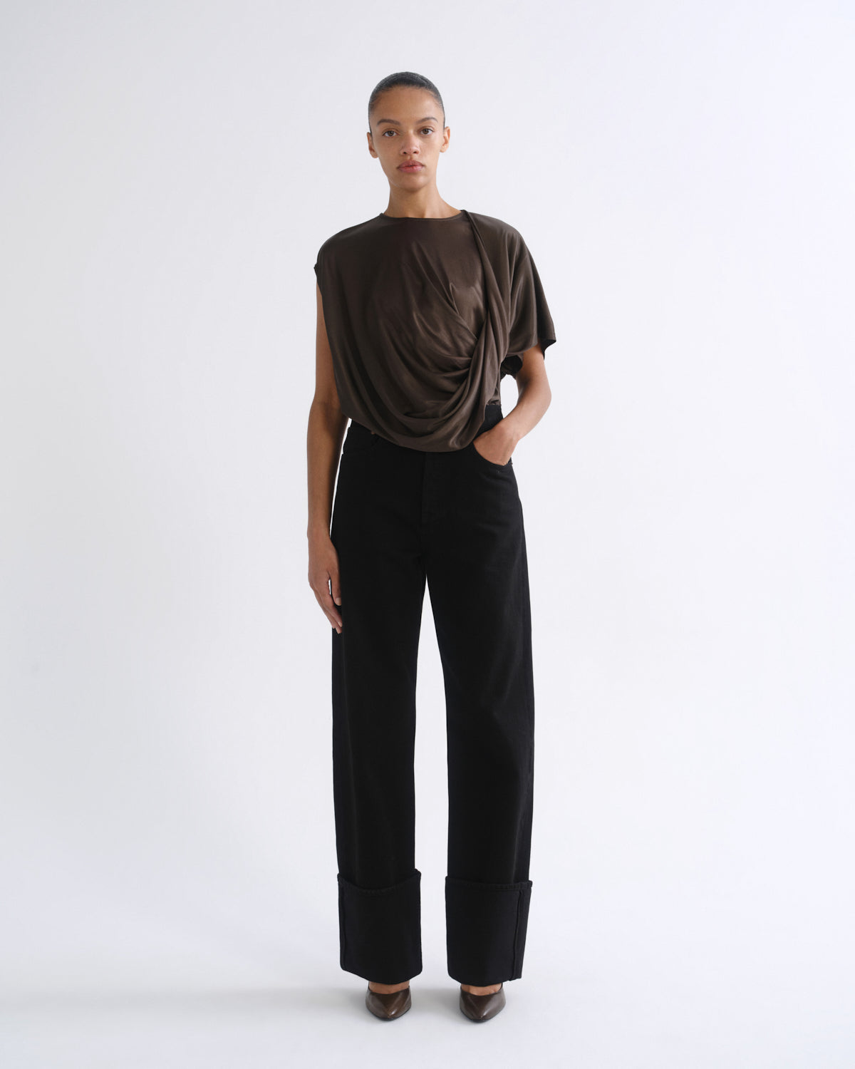A model stands against a plain white studio background wearing a dark brown draped short-sleeve top and black wide-leg trousers with rolled cuffs.