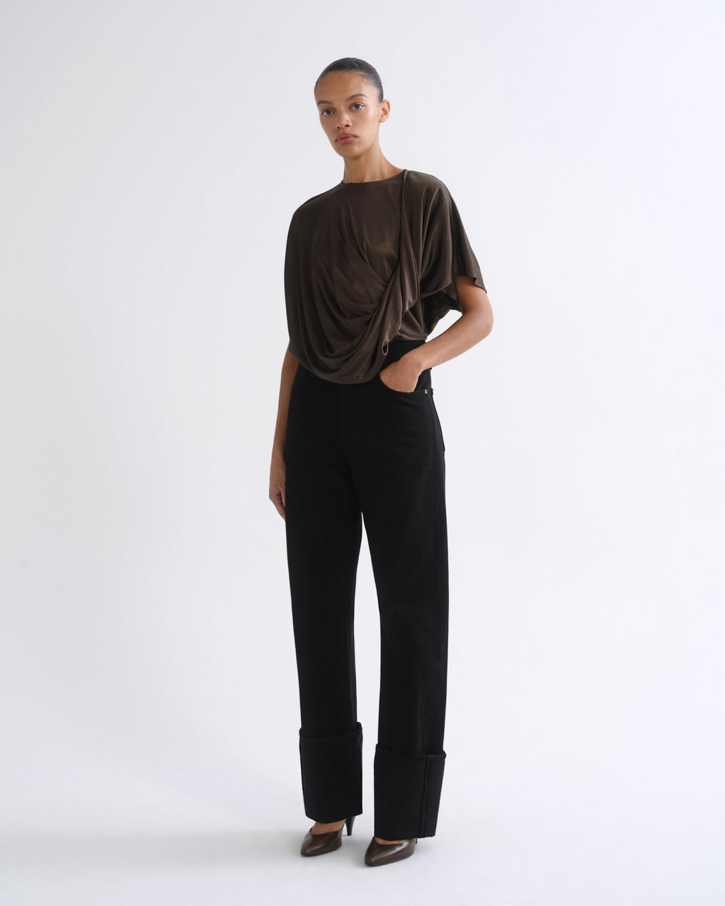 A model stands against a plain white studio background wearing a dark brown draped short-sleeve top and black wide-leg trousers with rolled cuffs, paired with brown pointed-toe heels.