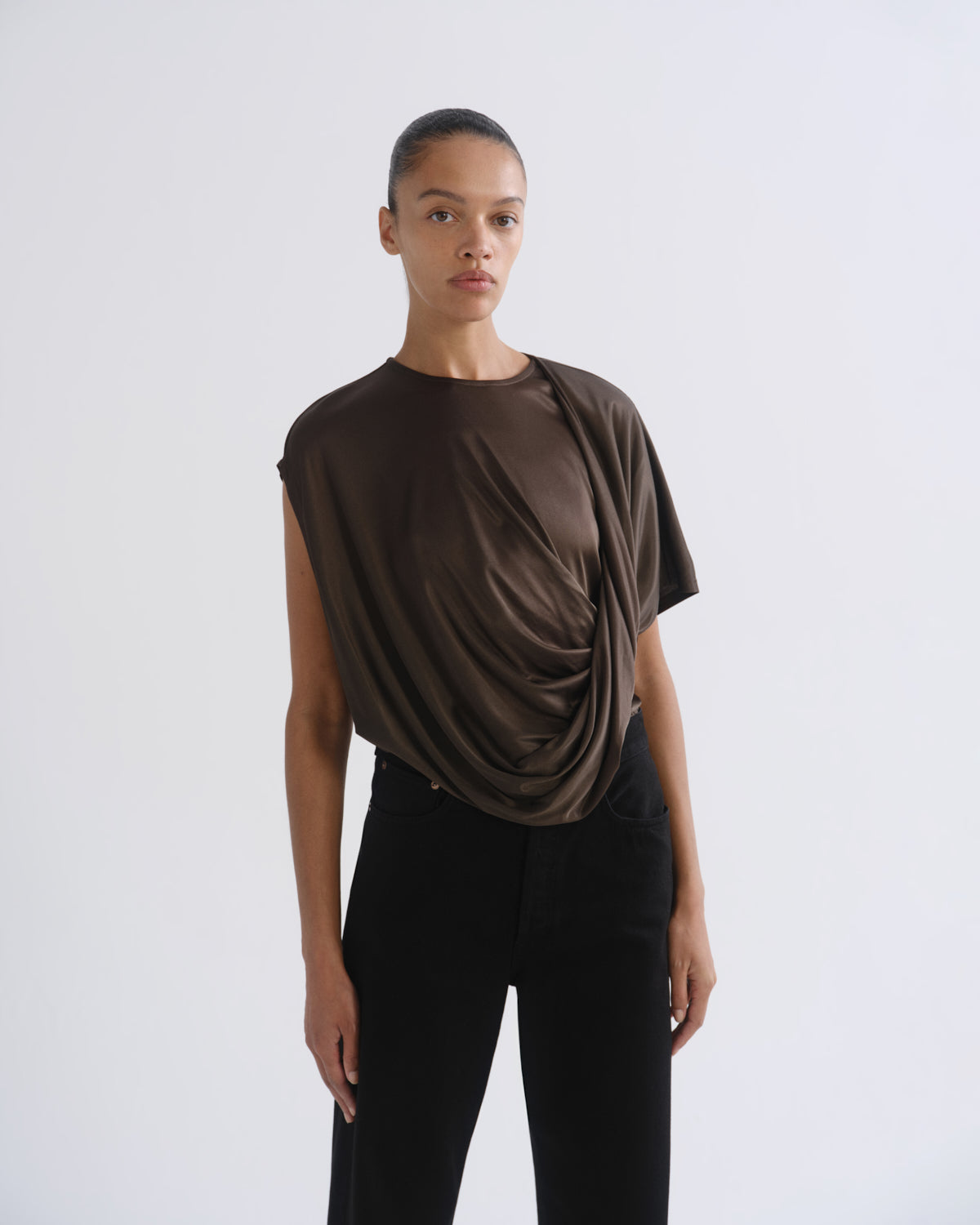 A model wearing a dark brown draped short-sleeve top and black pants, standing against a plain light gray studio background.