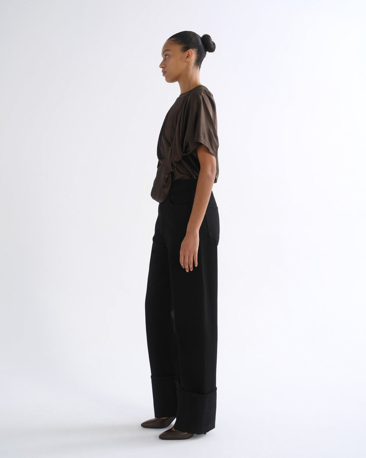 A model stands in profile wearing a dark brown short-sleeve top with gathered shoulders and black wide-leg trousers, photographed against a clean white studio background.