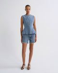 A model stands against a light gray studio background wearing a sleeveless light blue denim peplum top and matching denim shorts with frayed hems, paired with black strappy heels.