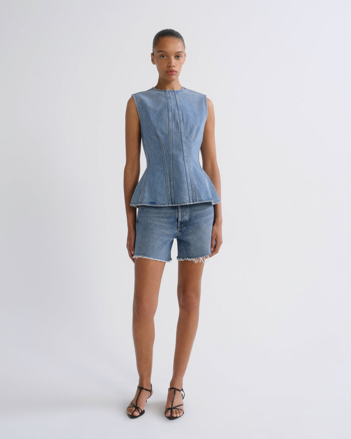 A model stands against a light gray studio background wearing a sleeveless light blue denim peplum top and matching denim shorts with frayed hems, paired with black strappy heels.