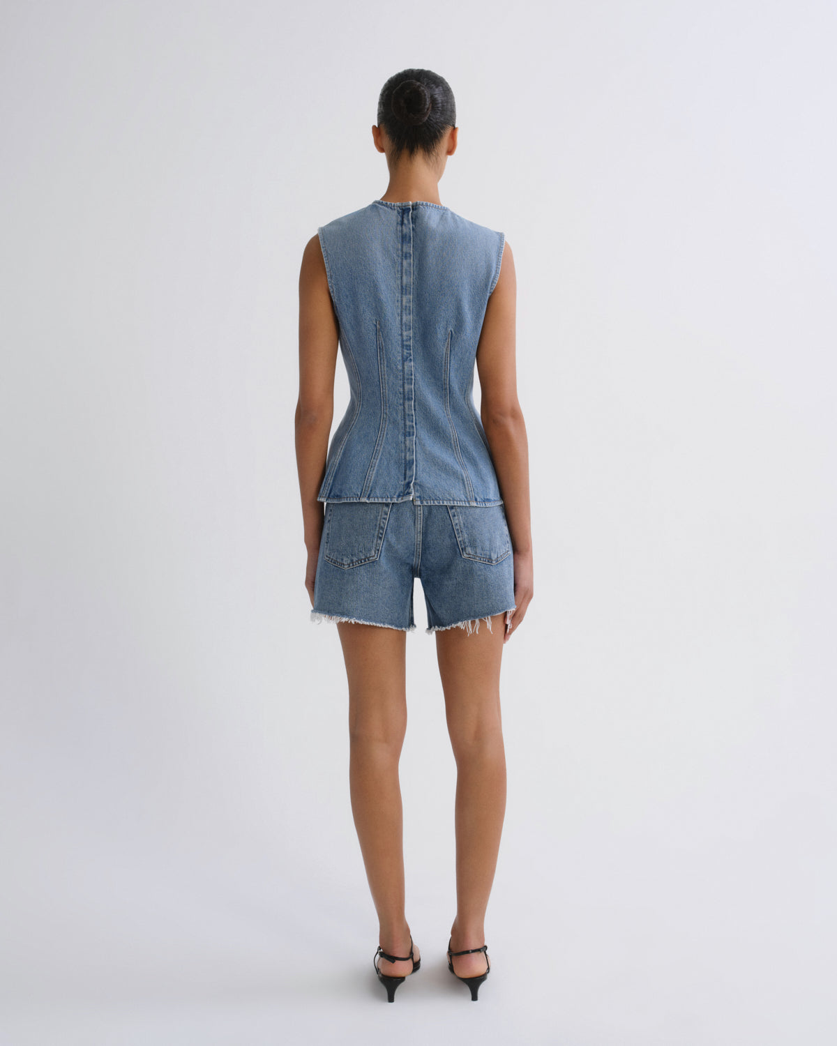 A model viewed from the back wearing a sleeveless light blue denim vest and matching denim shorts with frayed hems, paired with black high heels, against a plain light gray studio background.