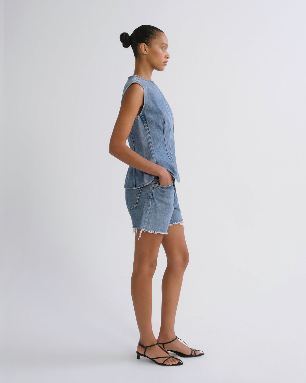 A model is shown in profile wearing a light blue denim sleeveless top and matching denim shorts with raw hems, paired with black strappy sandals, against a plain light gray studio background.