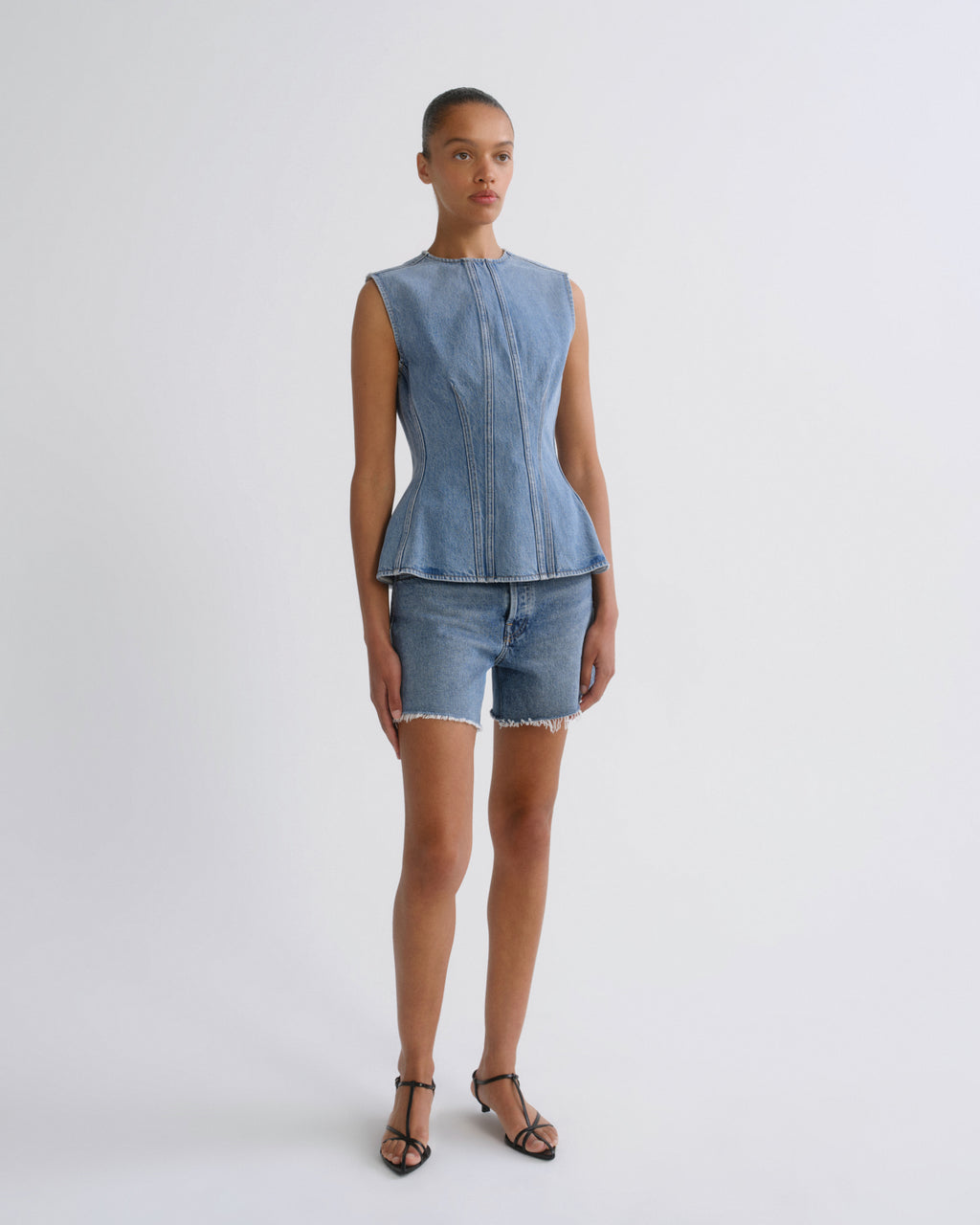 A model stands against a light gray studio background wearing a light blue denim sleeveless peplum top and matching denim shorts with fringe, paired with black strappy sandals.