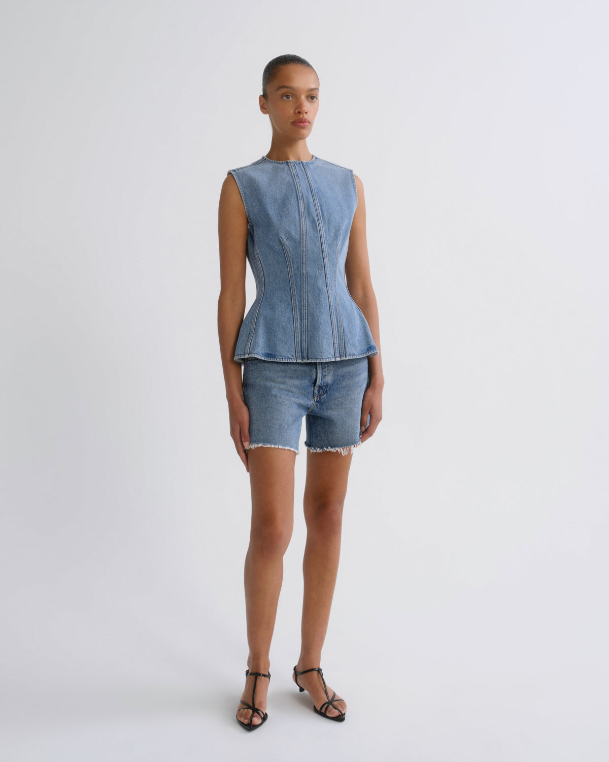 A model stands against a light gray studio background wearing a light blue denim sleeveless peplum top and matching denim shorts with fringe, paired with black strappy sandals.