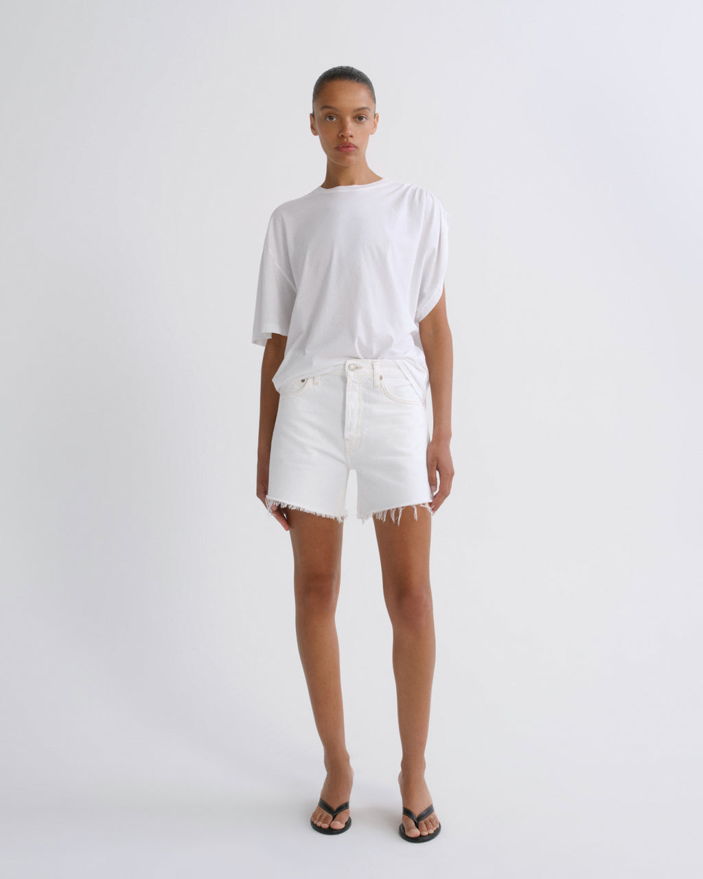 A model stands against a plain light gray studio background wearing a loose white short-sleeve T-shirt, white frayed-hem shorts, and black flip-flop sandals.