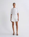 A model stands against a plain light gray studio background wearing a loose white short-sleeve T-shirt, white frayed-hem shorts, and black flip-flop sandals.