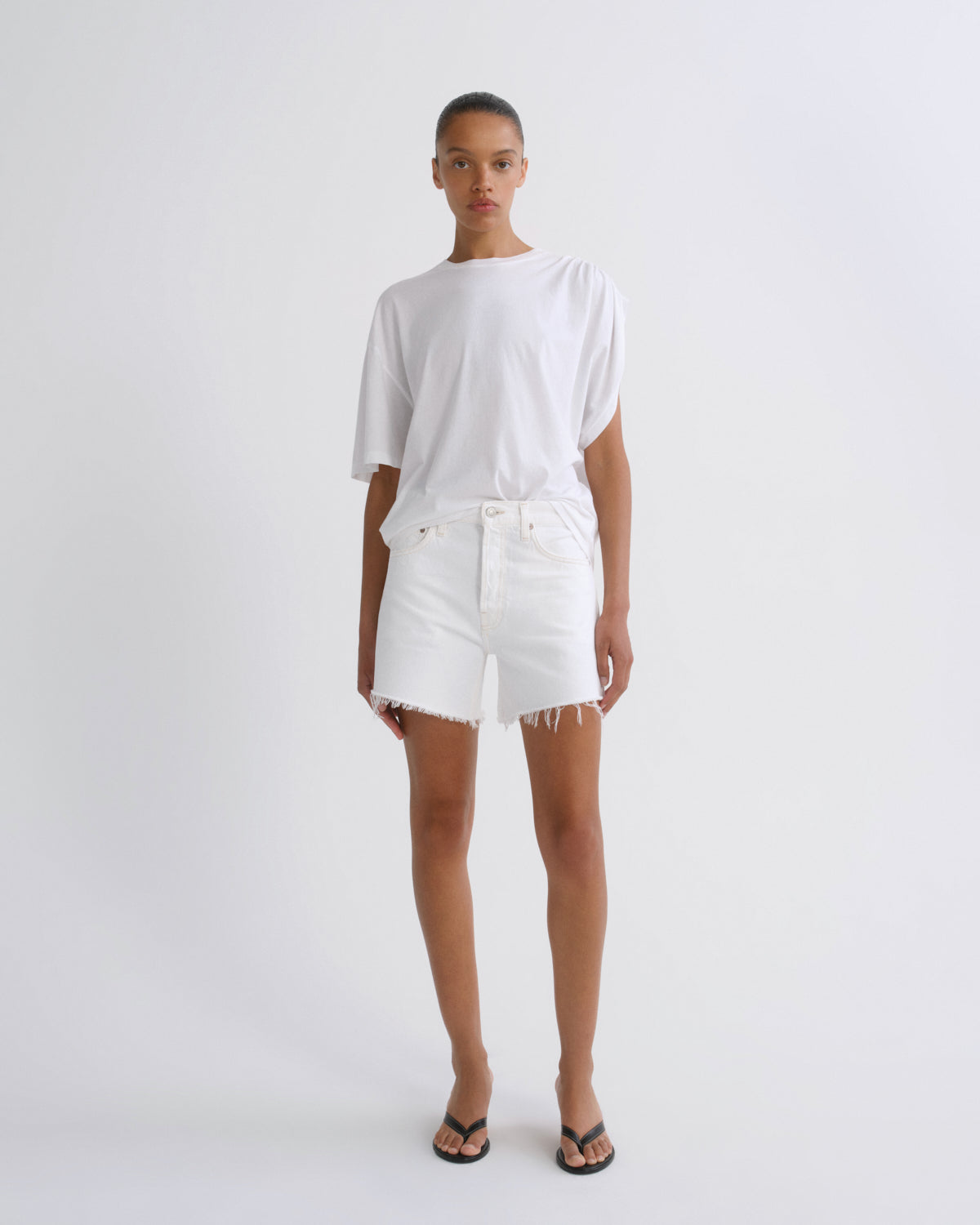 A model stands against a plain light gray studio background wearing a loose white short-sleeve T-shirt, white frayed-hem shorts, and black flip-flop sandals.