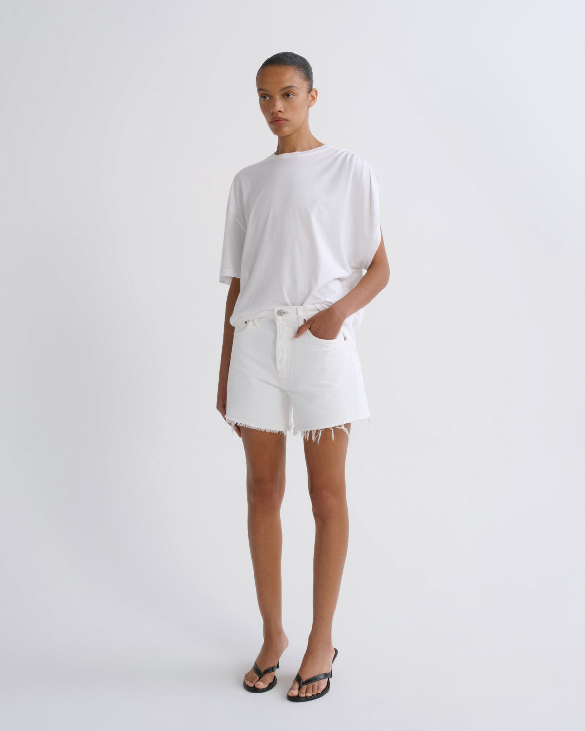 A model stands against a plain light gray studio background wearing a loose white short-sleeve T-shirt, white shorts with a frayed hem, and black flip-flop sandals.