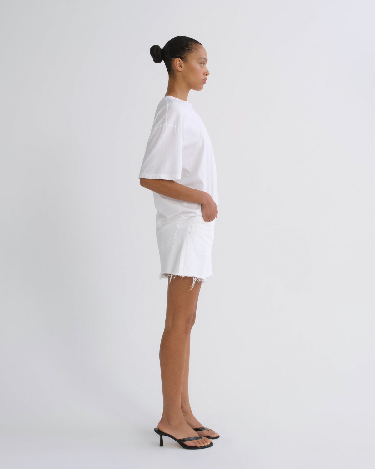 A model in profile wearing a white oversized short-sleeve top and matching white shorts with fringe, paired with black heeled sandals, against a plain light gray studio background.